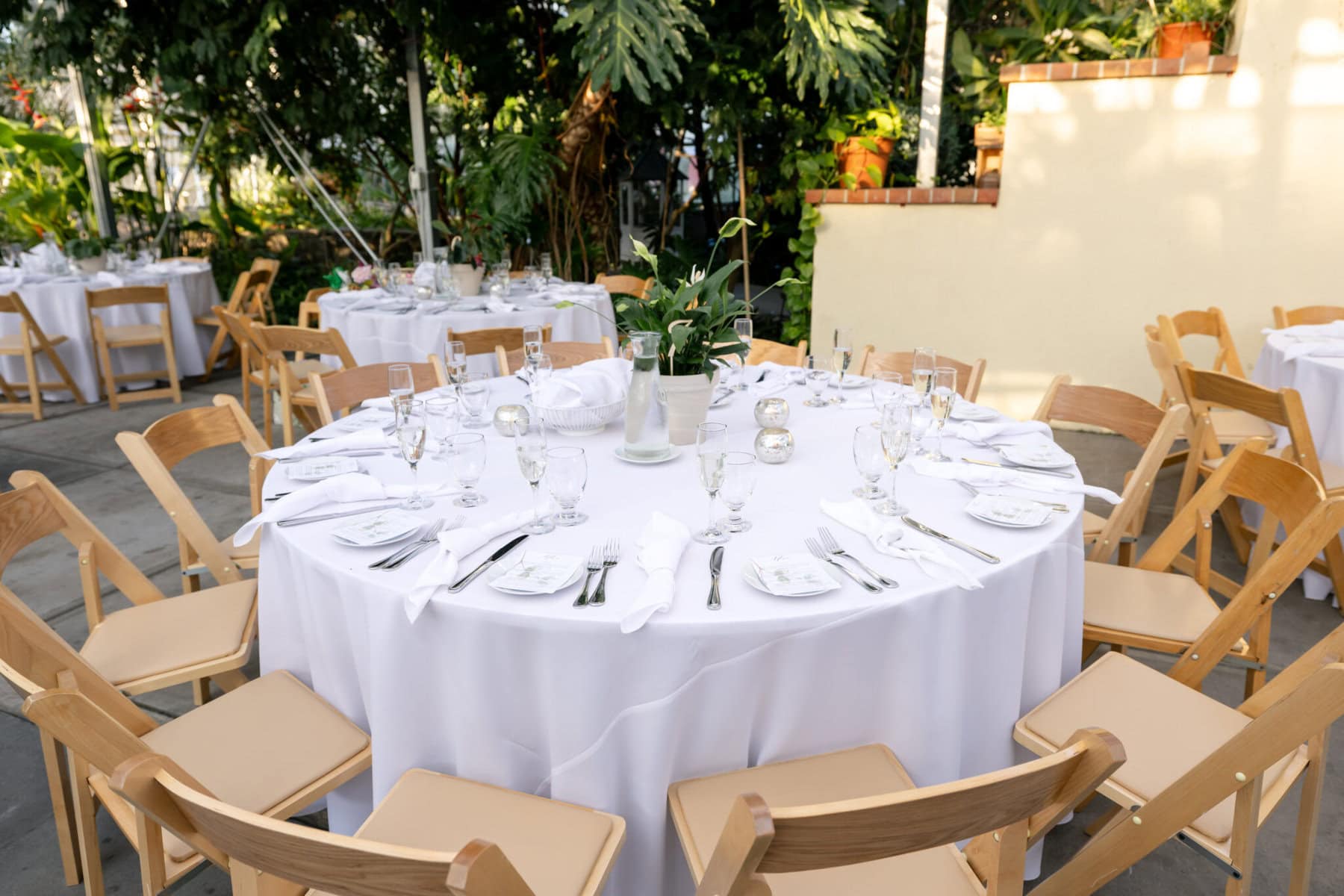 Stunning Wedding at the Roger Williams Botanical Gardens 61 Round table set with white linens and cutlery, surrounded by wooden chairs for a wedding at Roger Williams Botanical Gardens.