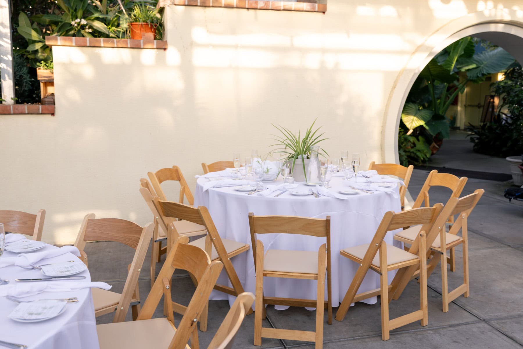 Stunning Wedding at the Roger Williams Botanical Gardens 62 A round table with a white tablecloth, set for a wedding at Roger Williams Botanical Gardens, surrounded by wooden chairs outdoors.