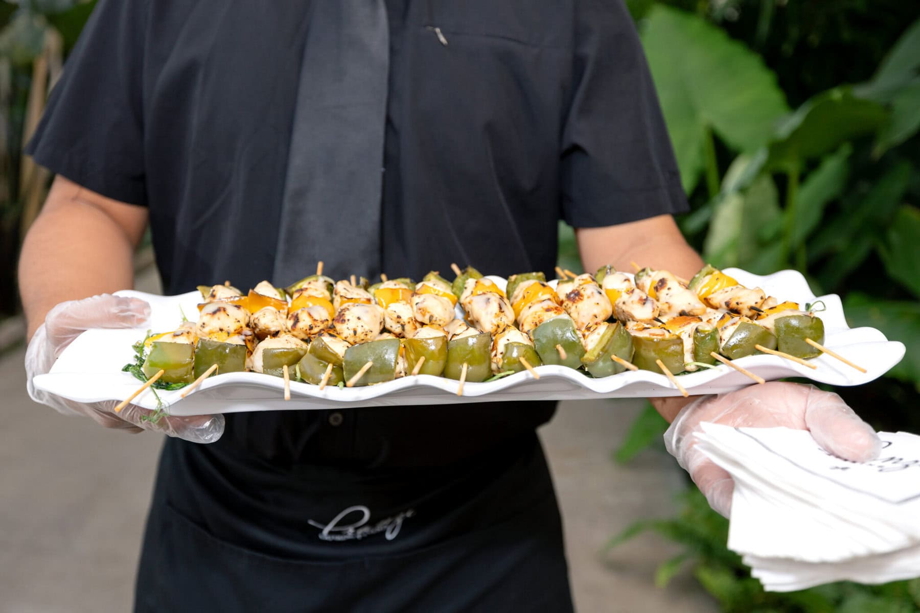 Stunning Wedding at the Roger Williams Botanical Gardens 45 A person in black clothing and gloves holds a tray of grilled chicken skewers at an outdoor wedding in Roger Williams Botanical Gardens.