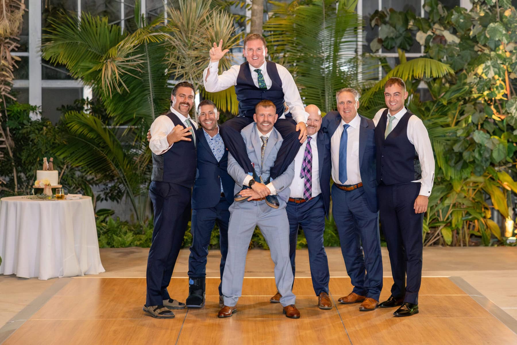 Stunning Wedding at the Roger Williams Botanical Gardens 78 Seven men in suits pose for a wedding photo at Roger Williams Botanical Gardens, with green plants behind and one man on another’s shoulders.