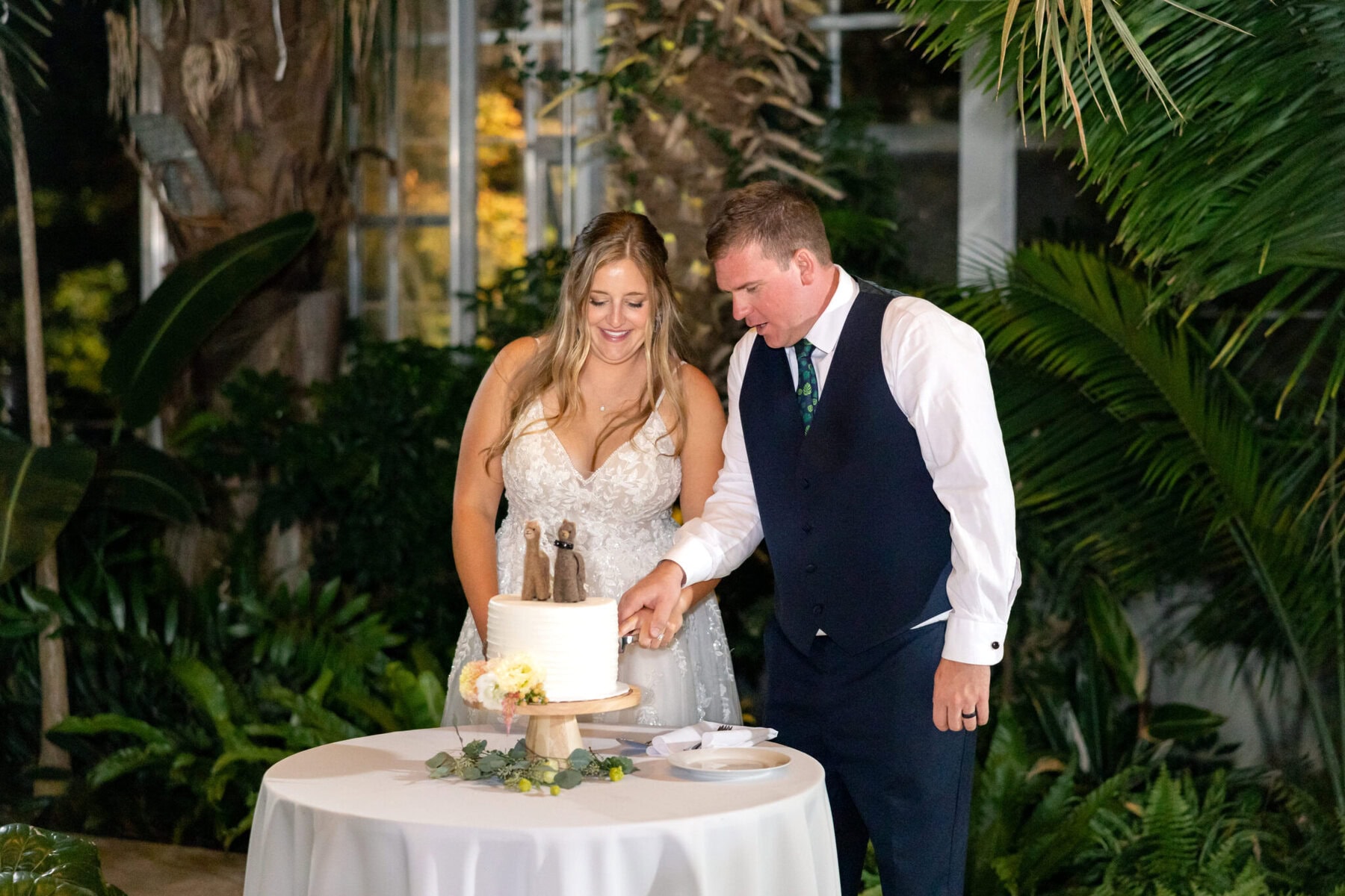 Stunning Wedding at the Roger Williams Botanical Gardens 80 A couple cuts their wedding cake together at a table, surrounded by lush green plants at Roger Williams Botanical Gardens.