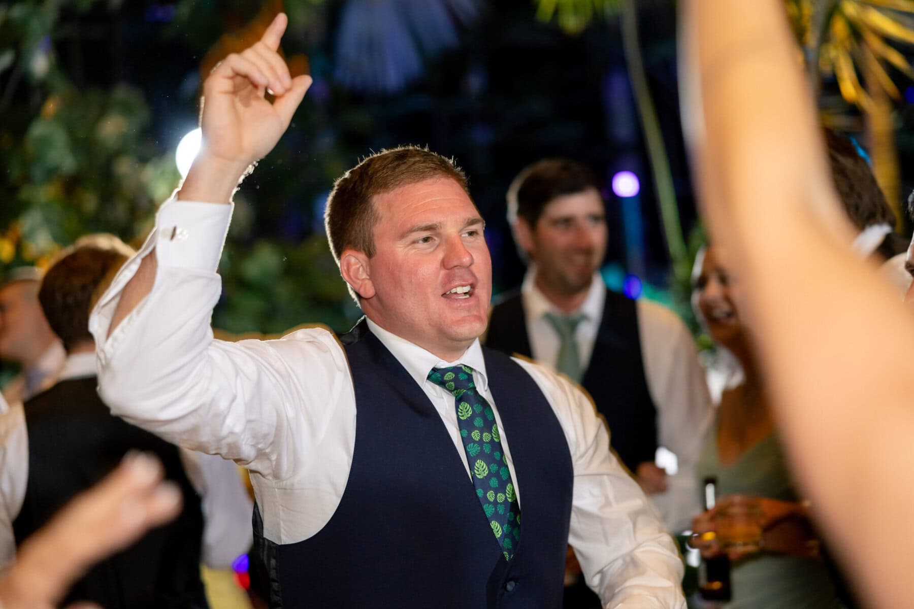 Stunning Wedding at the Roger Williams Botanical Gardens 82 A man in a suit vest and tie raises his hand while dancing at a wedding at Roger Williams Botanical Gardens, surrounded by formal guests.
