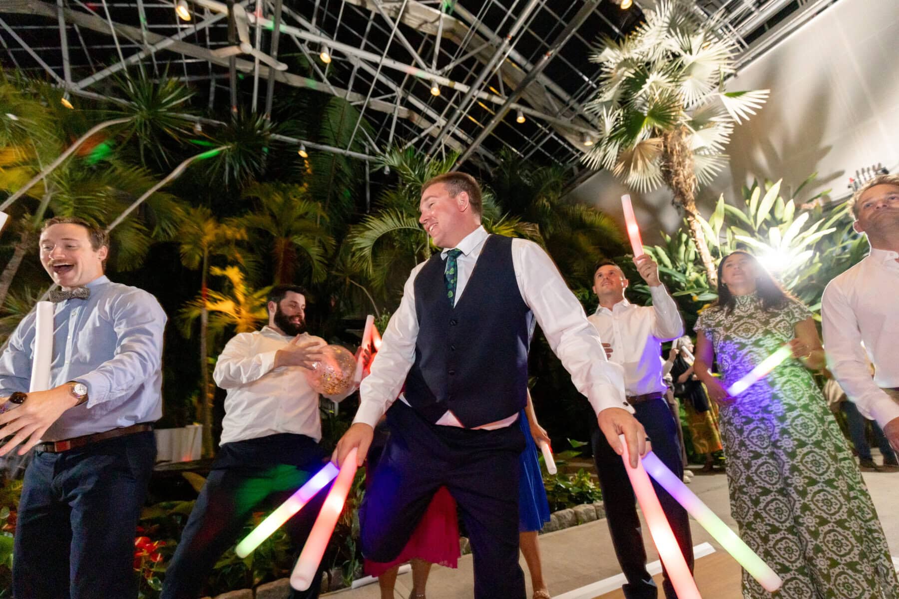 Stunning Wedding at the Roger Williams Botanical Gardens 83 Guests in formal attire dance with glowing light sticks at a wedding amid tropical plants at Roger Williams Botanical Gardens.