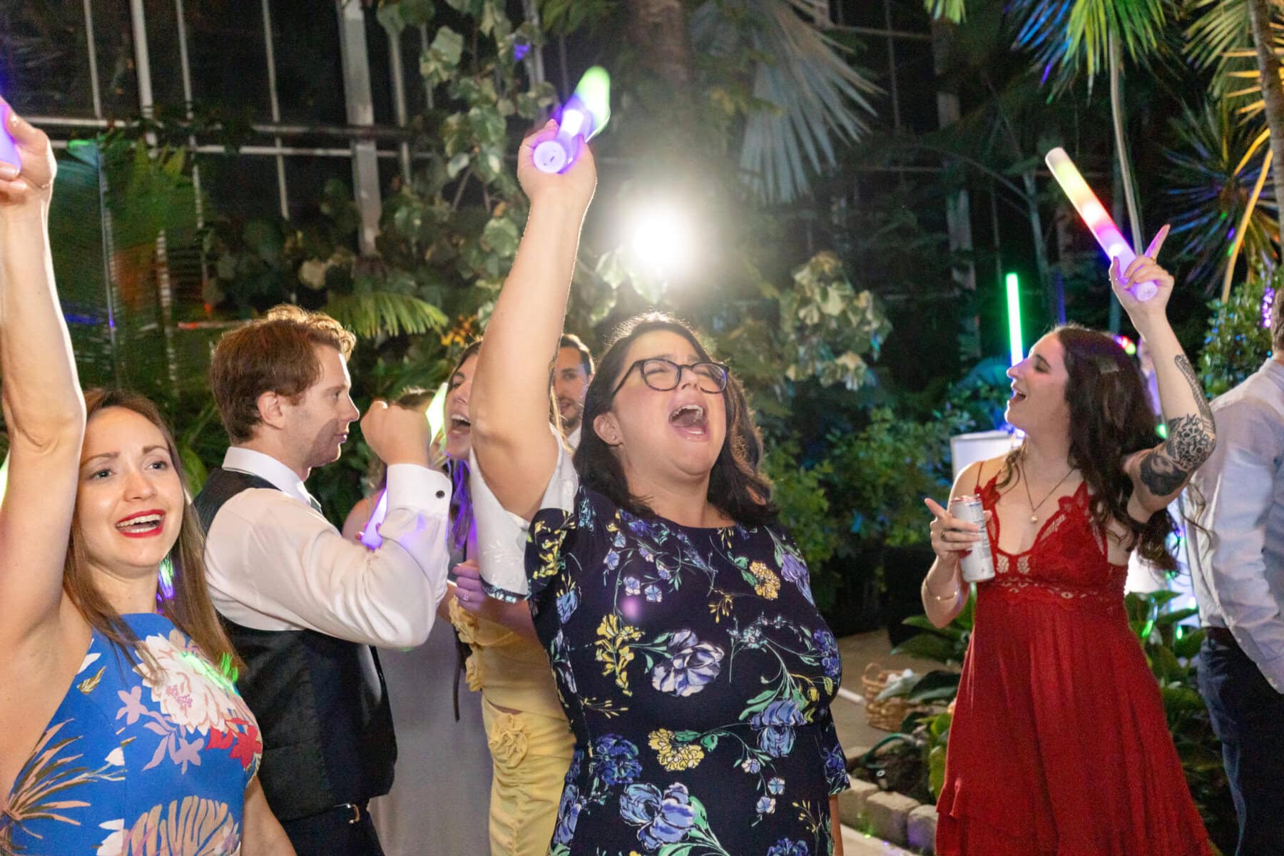 Stunning Wedding at the Roger Williams Botanical Gardens 85 People are dancing and cheering with glow sticks at a wedding in Roger Williams Botanical Gardens, surrounded by plants and colorful lights.