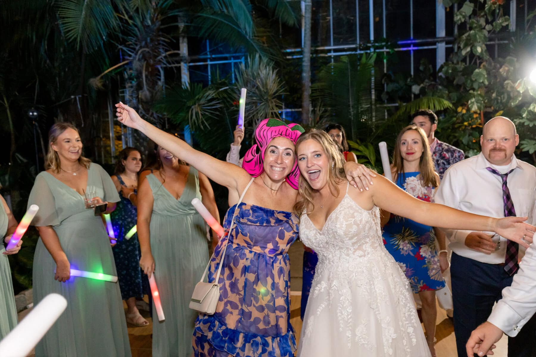 Stunning Wedding at the Roger Williams Botanical Gardens 87 A bride and a guest with arms raised smile at a wedding; other guests with glow sticks stand around them in festive attire.