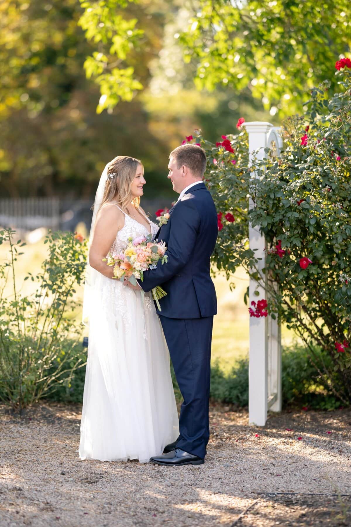 Stunning Wedding at the Roger Williams Botanical Gardens 96 A bride and groom stand facing each other at their wedding, surrounded by greenery and blooming red roses at Roger Williams Botanical Gardens.