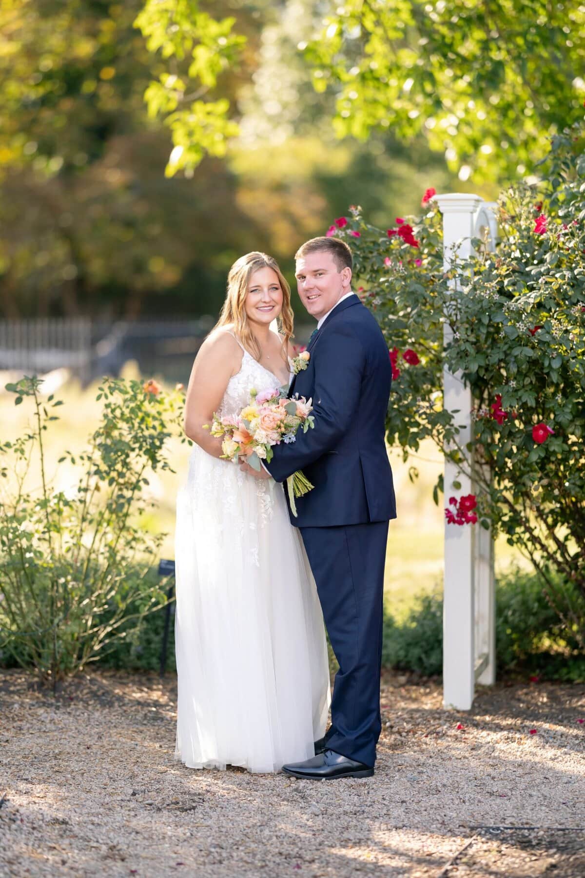 Stunning Wedding at the Roger Williams Botanical Gardens 97 A bride in a white dress and groom in a navy suit share a wedding moment outdoors at Roger Williams Botanical Gardens, bouquet in hand.