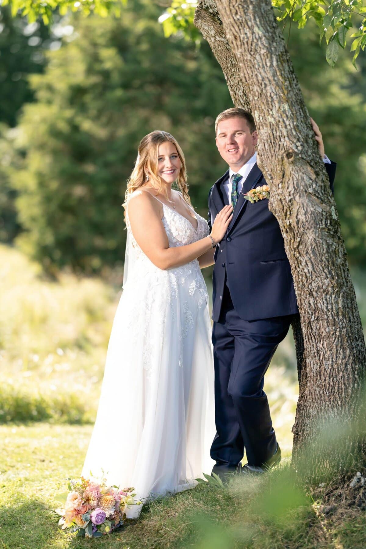 Stunning Wedding at the Roger Williams Botanical Gardens 99 A bride and groom pose by a tree at Roger Williams Botanical Gardens, with a wedding bouquet resting on the grass in front of them.