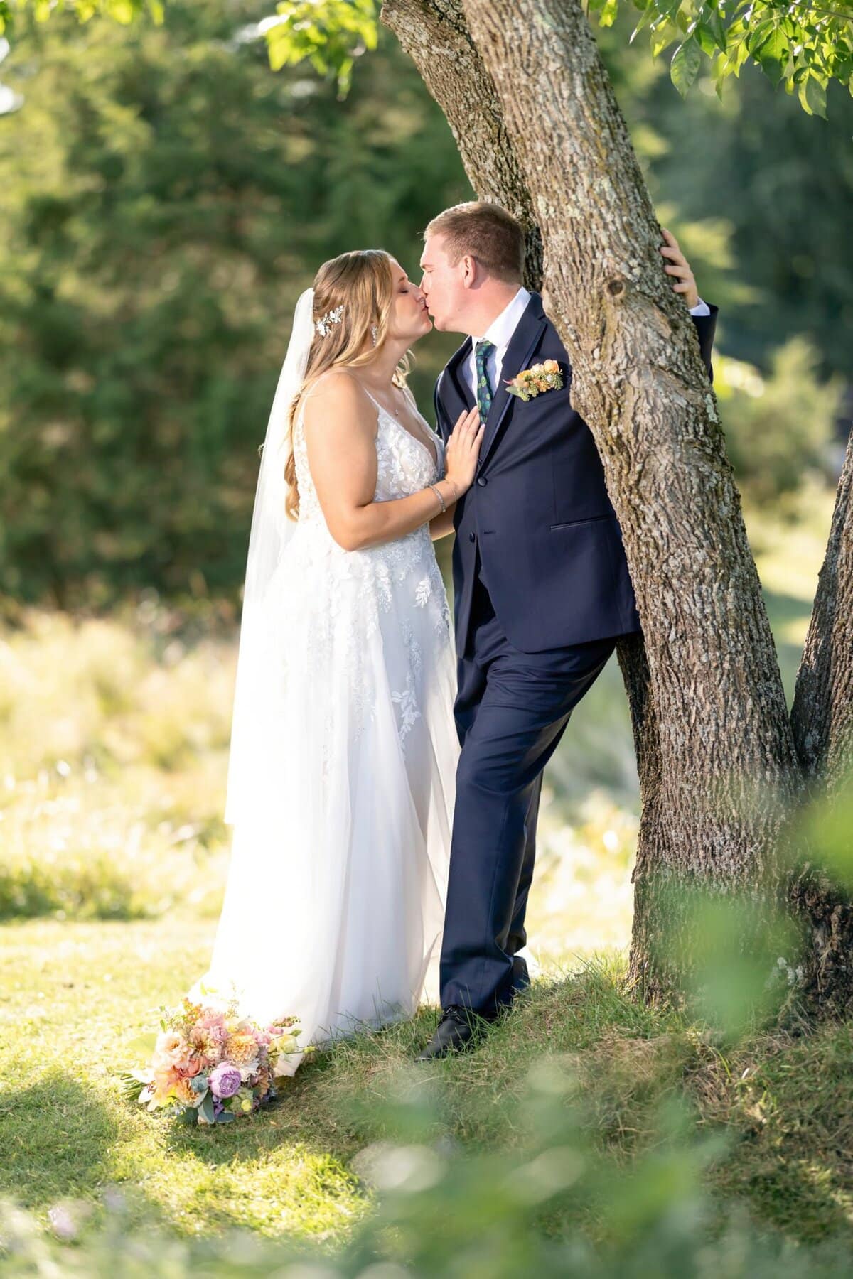 Stunning Wedding at the Roger Williams Botanical Gardens 100 A bride and groom share a kiss by a tree at Roger Williams Botanical Gardens; their bouquet rests on the grass nearby.