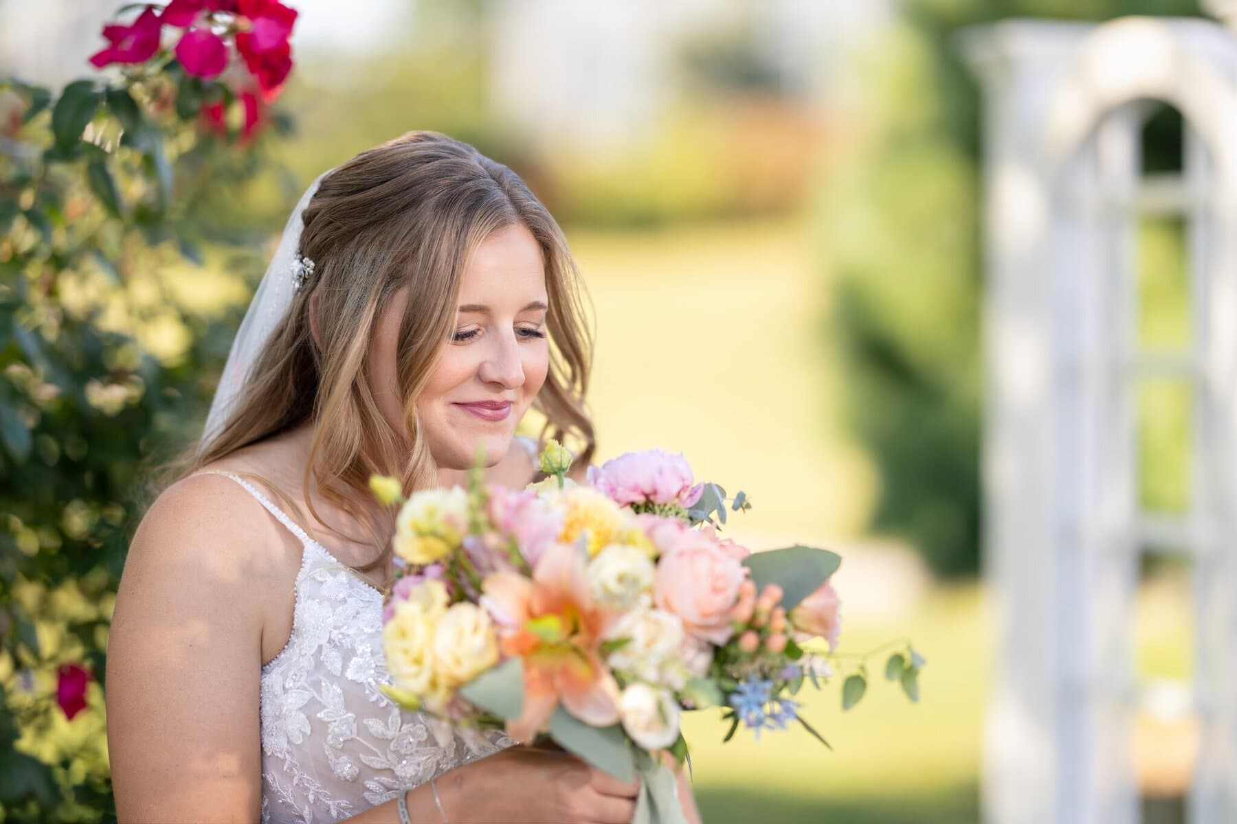 Stunning Wedding at the Roger Williams Botanical Gardens 94 A woman in a wedding dress smiles, holding a bouquet near greenery and a white structure at Roger Williams Botanical Gardens.