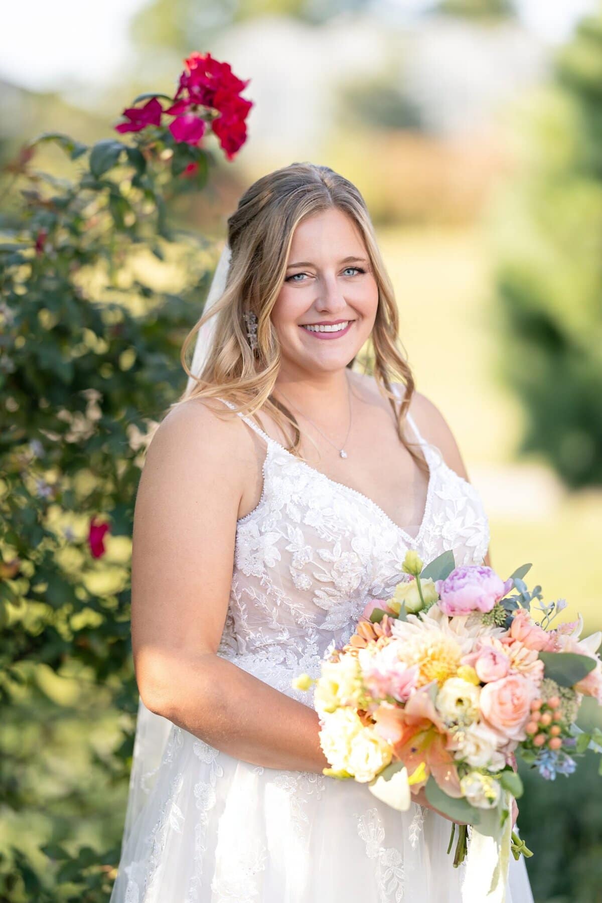 Stunning Wedding at the Roger Williams Botanical Gardens 95 A bride in a white lace dress smiles outdoors, holding her bouquet at the Roger Williams Botanical Gardens near vibrant red roses.