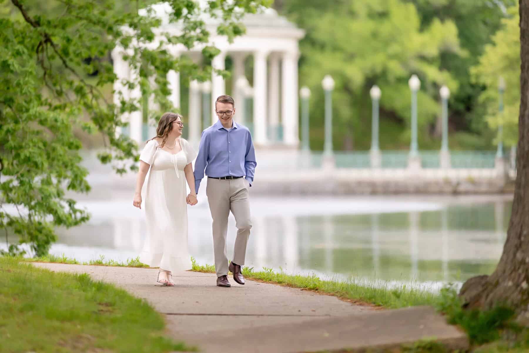 Roger Williams Park Engagement Session 31 A couple walks hand in hand on a lakeside path in Roger Williams Park, surrounded by green trees—a perfect engagement session with a white pavilion in the background.