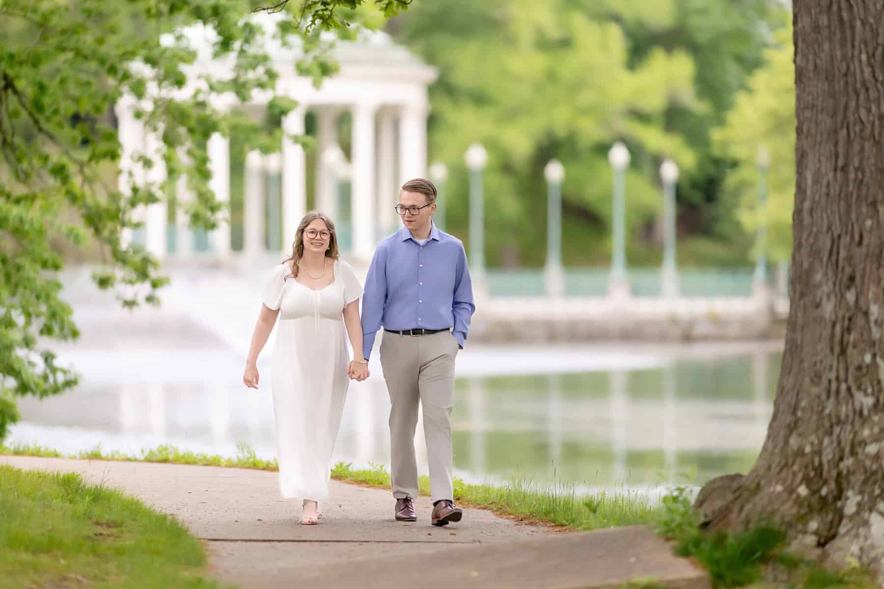 Roger Williams Park Engagement Session 18 A couple holds hands on a tree-lined path by a lake during their Roger Williams Park engagement session, with a white pavilion in the background.