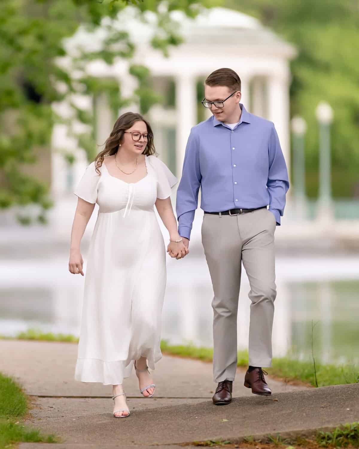 Roger Williams Park Engagement Session 21 A couple holding hands walks on a riverside path in Roger Williams Park. The woman wears a white dress; the man, a light blue shirt and khaki pants. Green trees and a white pavilion appear behind them.