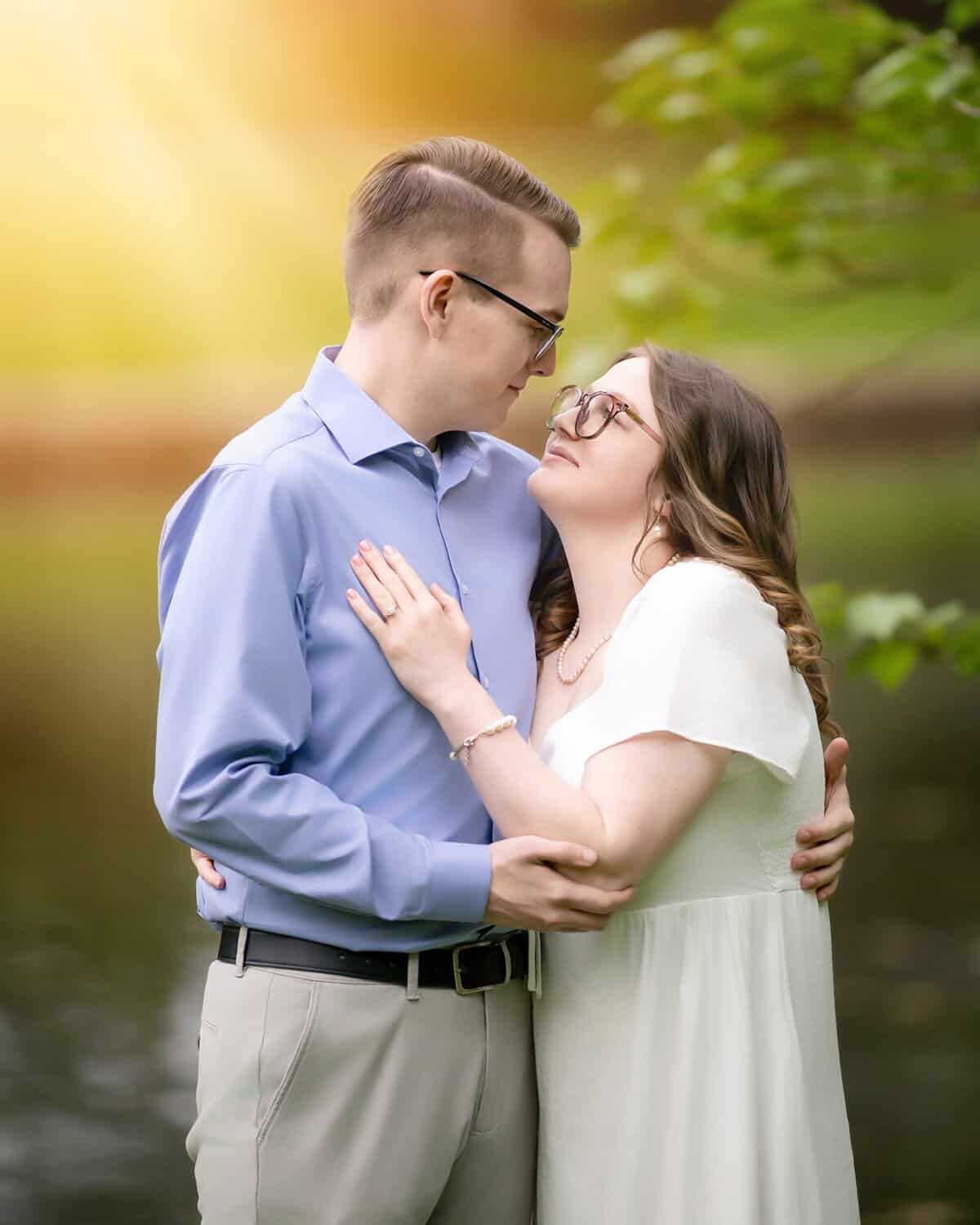 Roger Williams Park Engagement Session 22 A couple embraces by a calm body of water in Roger Williams Park, gazing into each other's eyes during their engagement session. Sunlight filters through green leaves above.