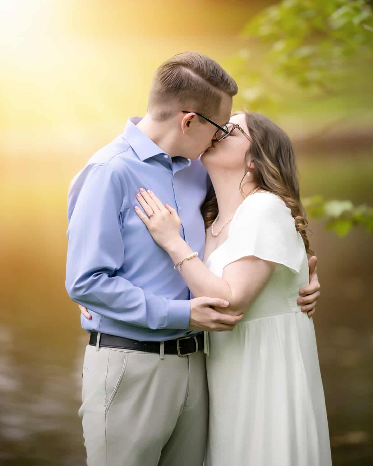 Roger Williams Park Engagement Session 23 A couple shares a kiss by a river in Roger Williams Park, embraced closely. Warm sunlight glows as greenery surrounds them during their engagement session.