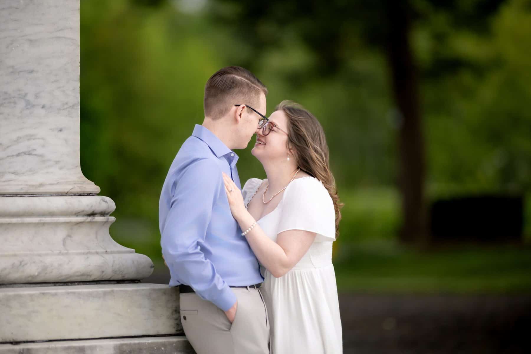 Roger Williams Park Engagement Session 26 A smiling couple stands close together outdoors by a marble pillar during their Roger Williams Park engagement session; she touches his chest as they look lovingly into each other’s eyes.