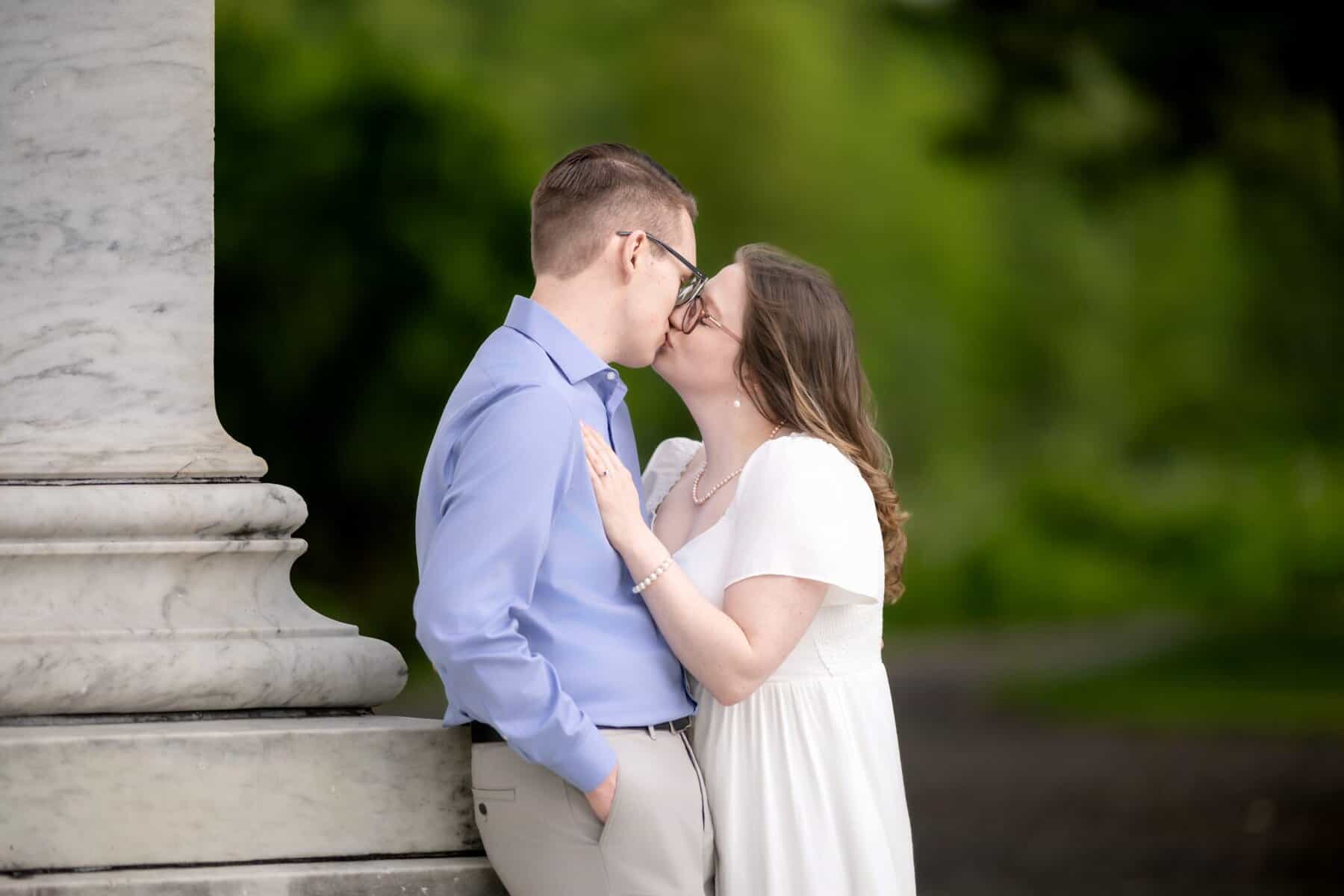 Roger Williams Park Engagement Session 27 A couple wearing glasses shares a kiss outdoors by a marble column during their engagement session at Roger Williams Park, with blurred green foliage in the background.