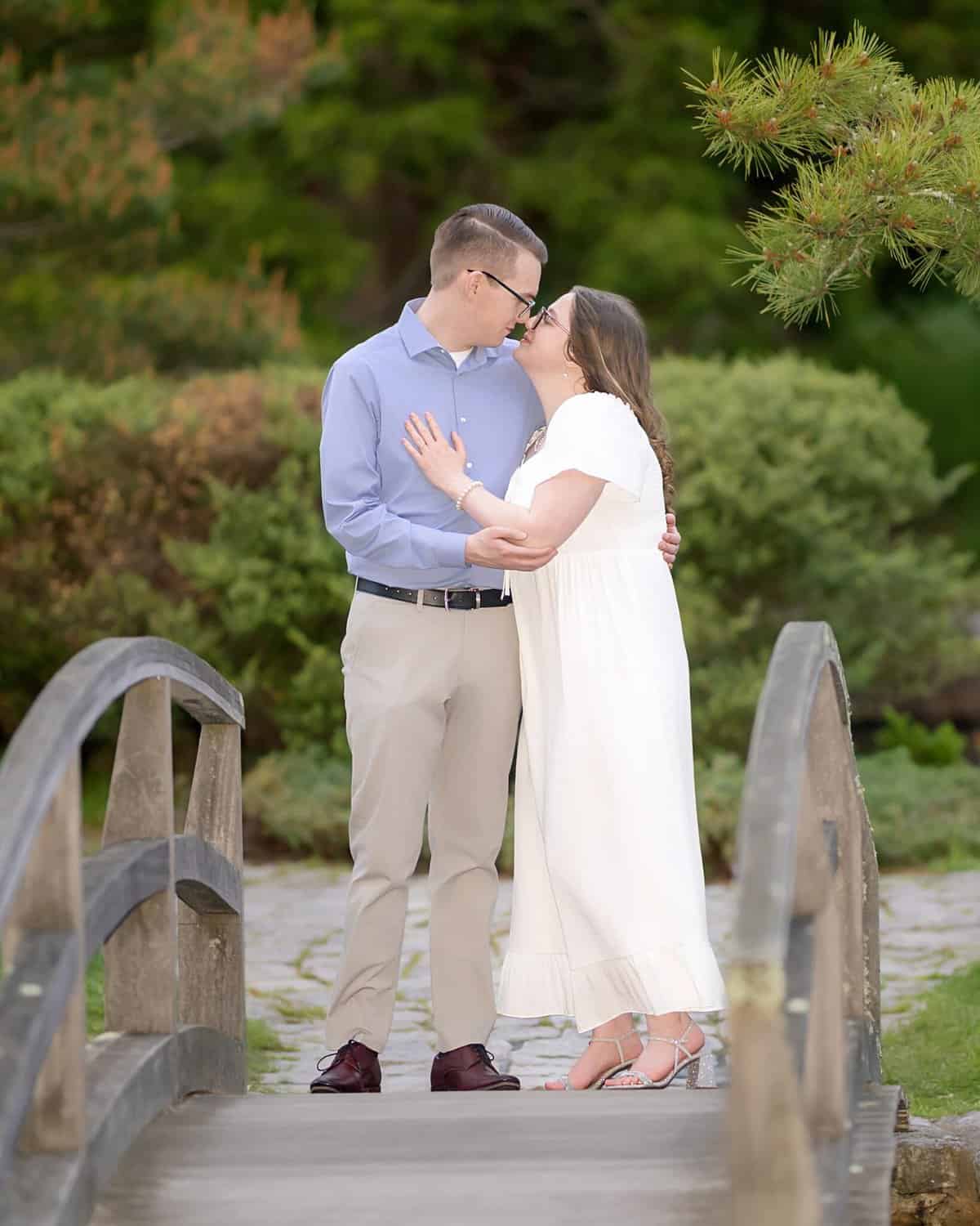 Roger Williams Park Engagement Session 2 A couple stands on a wooden bridge at Roger Williams Park, smiling and touching noses during their engagement session. Green trees and bushes fill the background.