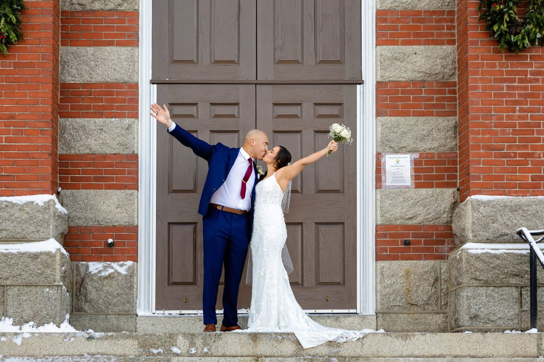 Wedding At the Transfiguration of the Lord Parish 54 A bride and groom celebrate and kiss on stone steps outside Transfiguration of the Lord Parish, with snowy touches around them.
