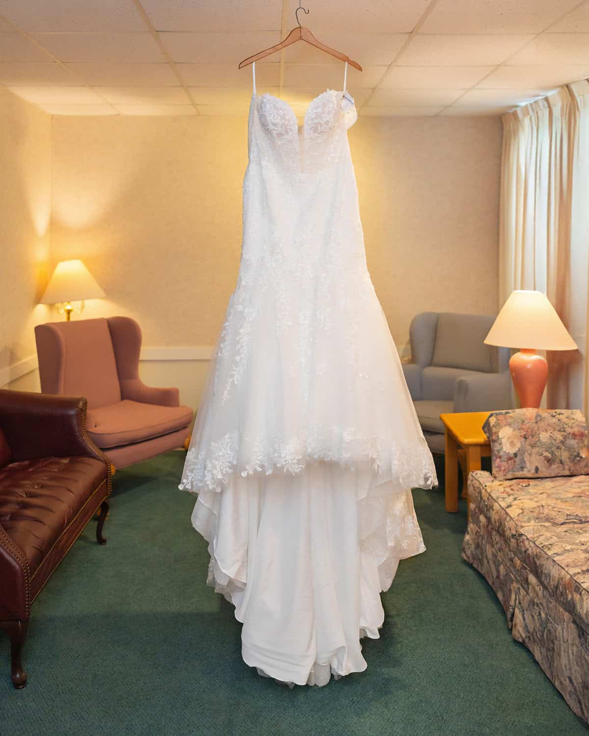Wedding at the Springfield Elks lodge 2 A white lace wedding dress hangs on a wooden hanger in a cozy room at the Springfield Elks Lodge, with pastel furniture, green carpet, and cream-colored walls and curtains.