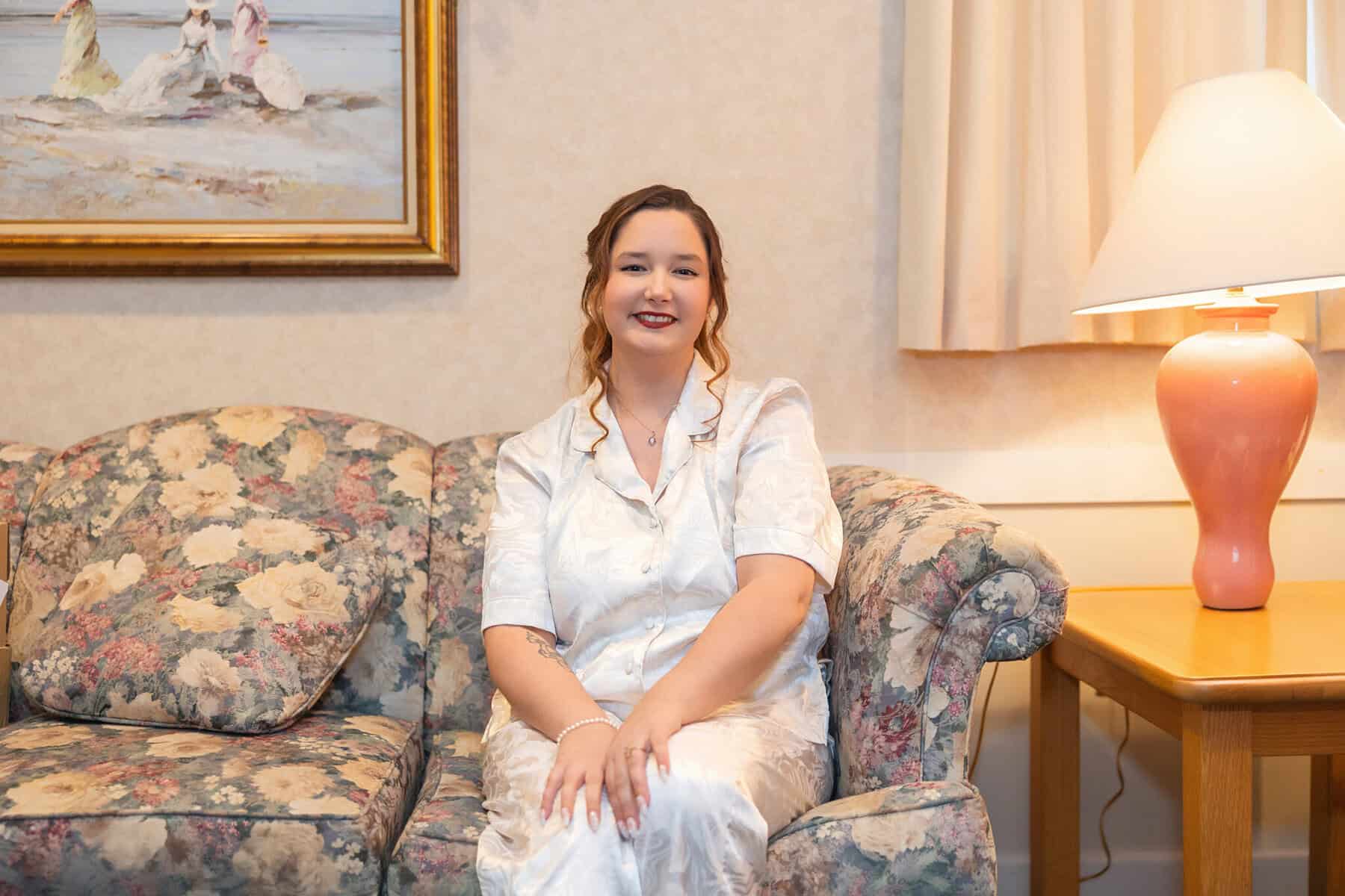 Wedding at the Springfield Elks lodge 3 A woman in white pajamas sits smiling on a floral couch in Springfield, hands folded, beside a pink lamp and painting—perhaps reflecting after an elks lodge wedding.