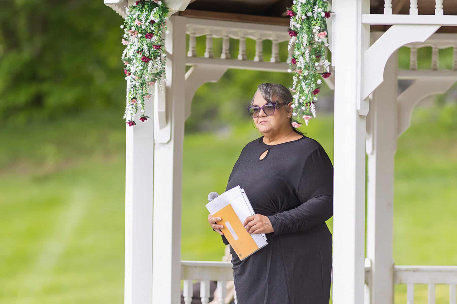 Wedding at the Springfield Elks lodge 6 A woman in sunglasses and a black dress stands in a white gazebo at an Elks Lodge wedding, holding papers beneath floral decorations, with Springfield's greenery blurred in the background.