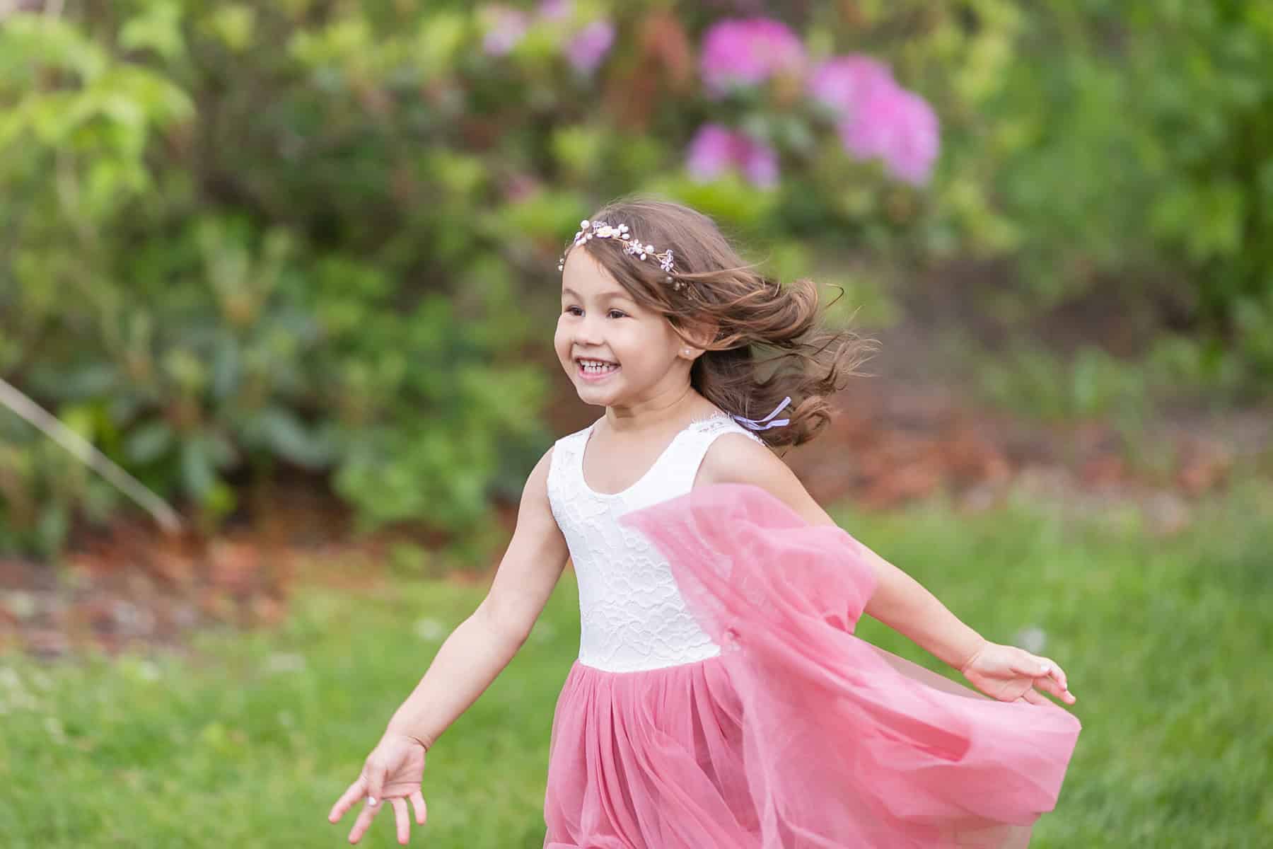 Wedding at the Springfield Elks lodge 7 A young girl in a pink and white dress runs joyfully on grass at a Springfield wedding, smiling. She wears a floral headband and her hair flows in the breeze amid blurred flowers.