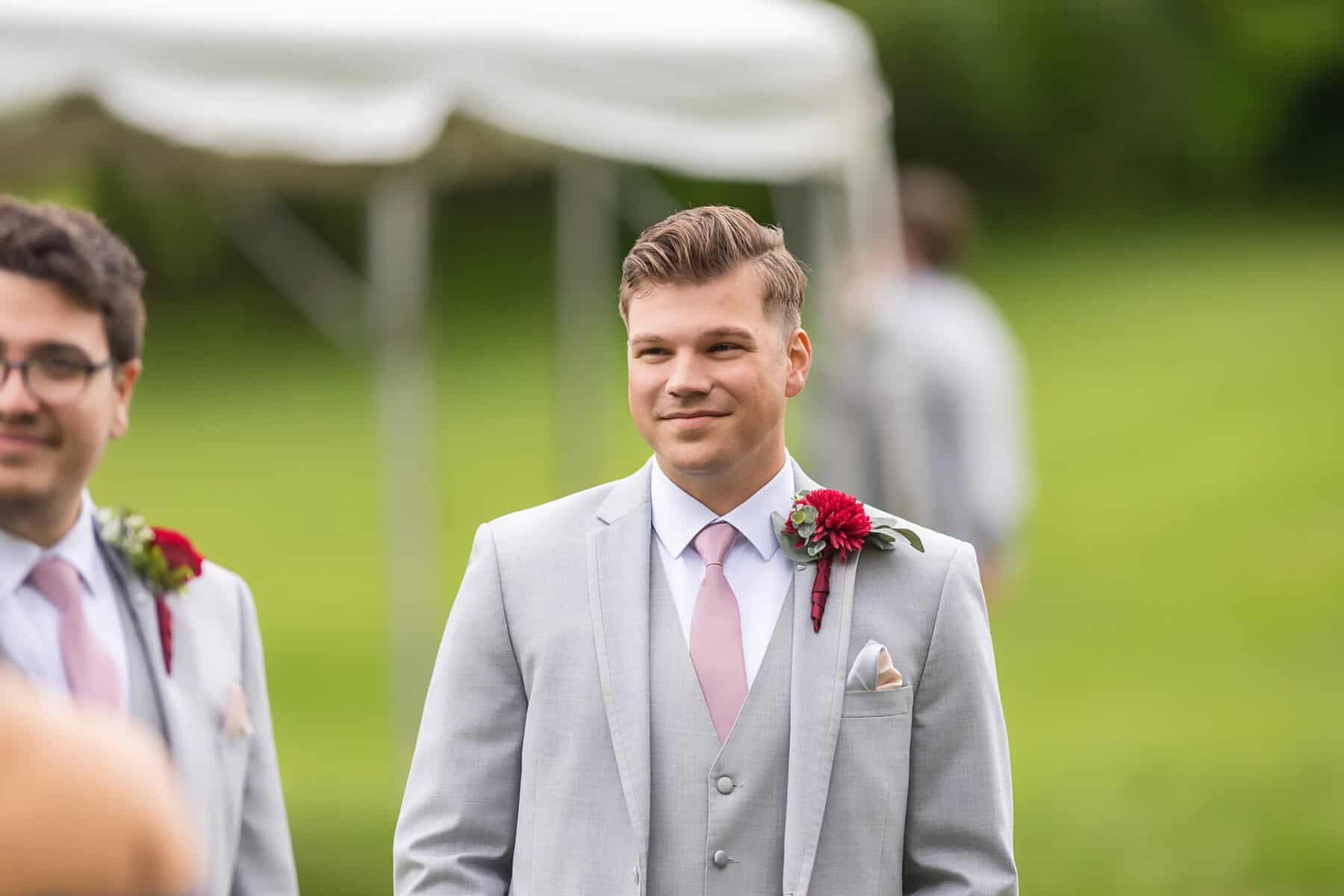 Wedding at the Springfield Elks lodge 8 A young man in a light gray suit with a pink tie and red boutonniere stands outdoors at a Springfield wedding, smiling, with blurred greenery and a white tent in the background.