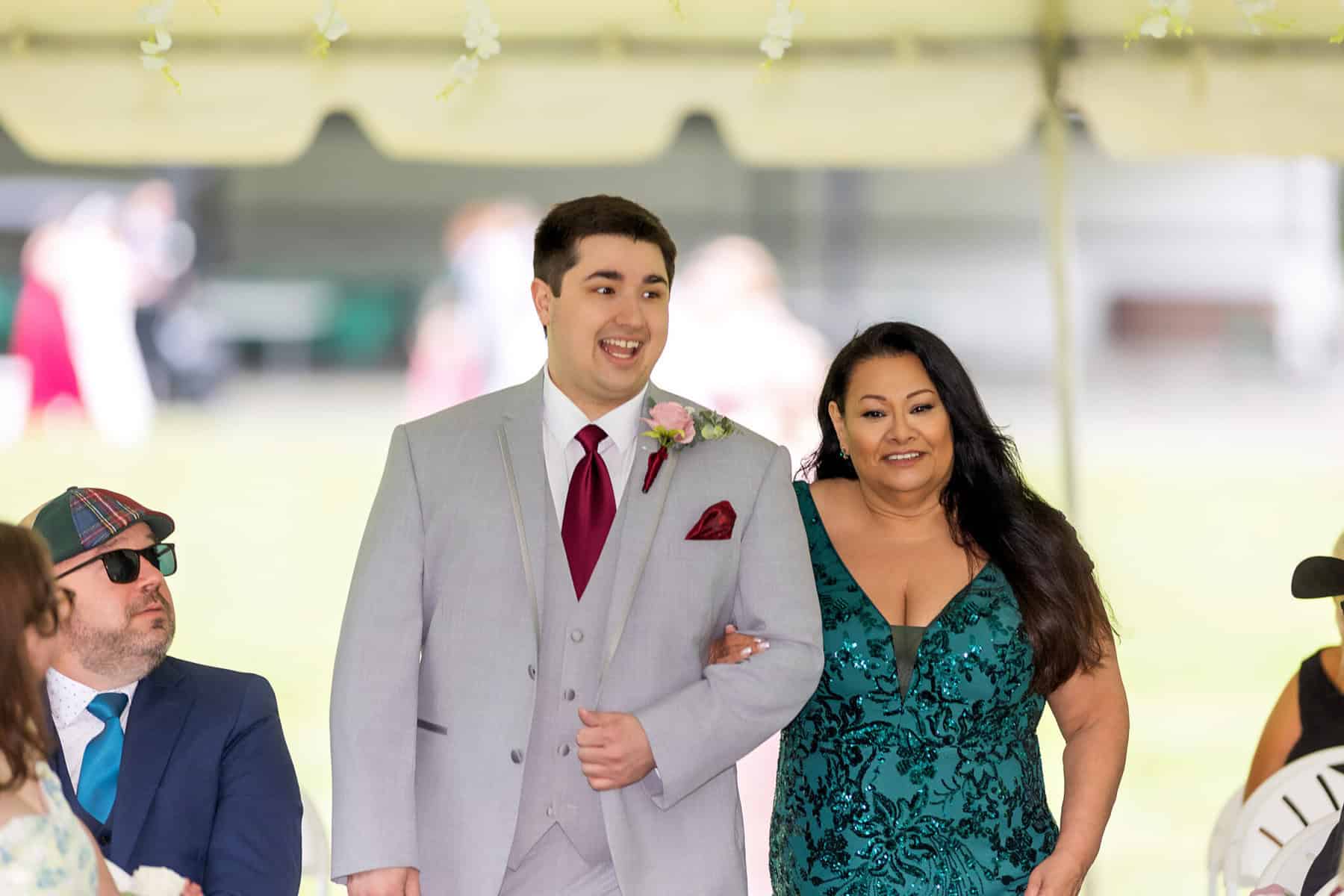 Wedding at the Springfield Elks lodge 11 A smiling man in a light gray suit with a burgundy tie walks arm-in-arm with a woman in a green dress at a Springfield wedding, guests seated around them at the outdoor Elks Lodge event.