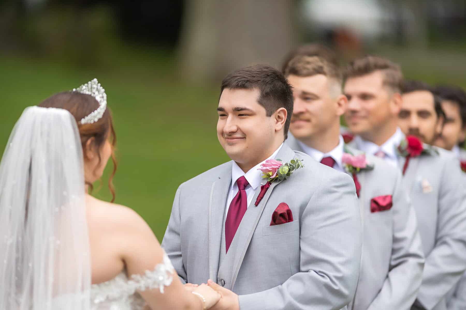Wedding at the Springfield Elks lodge 17 A groom in a light gray suit with a red tie smiles at his bride outdoors at the Springfield Elks Lodge, with groomsmen in matching suits lined up behind him on their wedding day.
