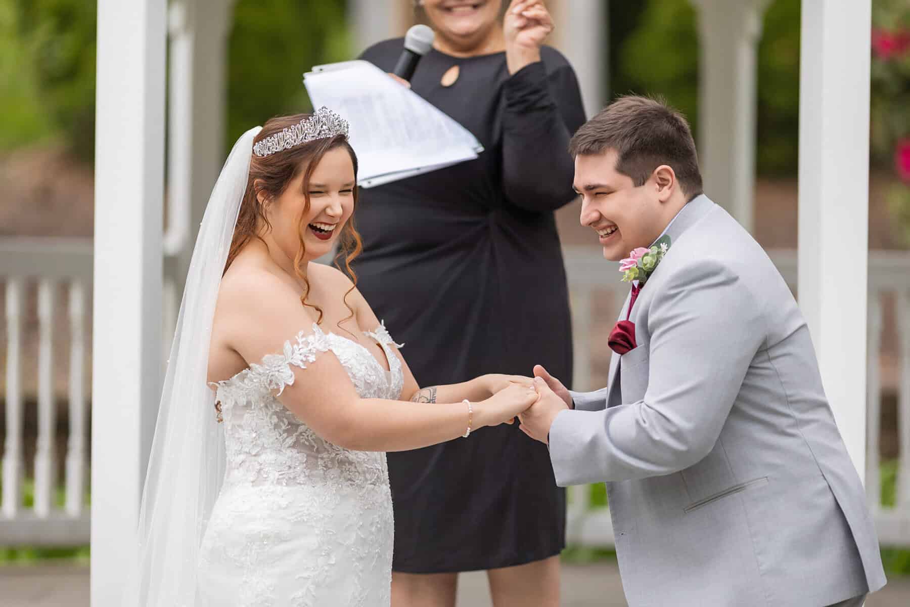 Wedding at the Springfield Elks lodge 21 A bride and groom laugh joyfully, holding hands during their outdoor wedding ceremony at the Elks Lodge in Springfield, while an officiant smiles behind them with a microphone and clipboard.