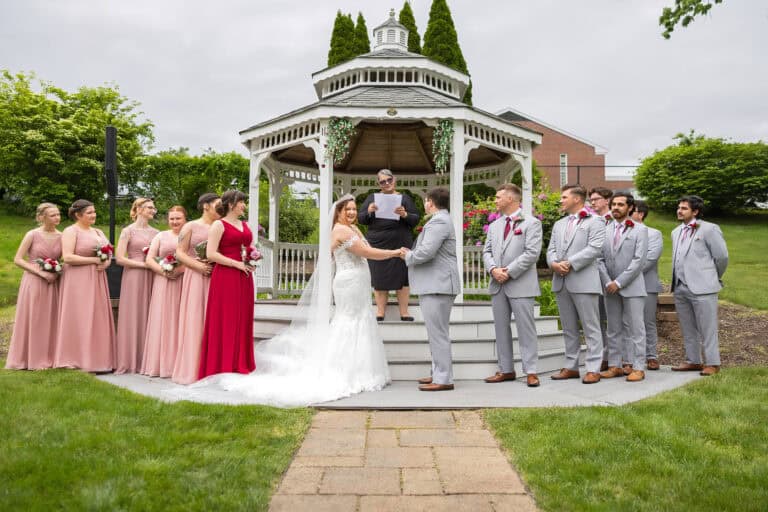 A bride and groom hold hands at an outdoor Elks Lodge wedding in Springfield, surrounded by bridesmaids in pink and groomsmen in gray, as the officiant stands behind them.