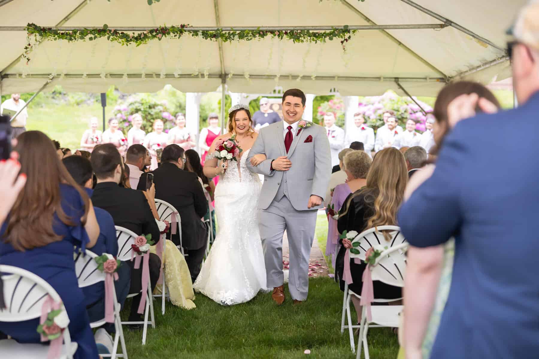 Wedding at the Springfield Elks lodge 26 A smiling bride and groom walk down the aisle under a decorated tent at the Springfield Elks Lodge, surrounded by guests capturing the wedding moment.
