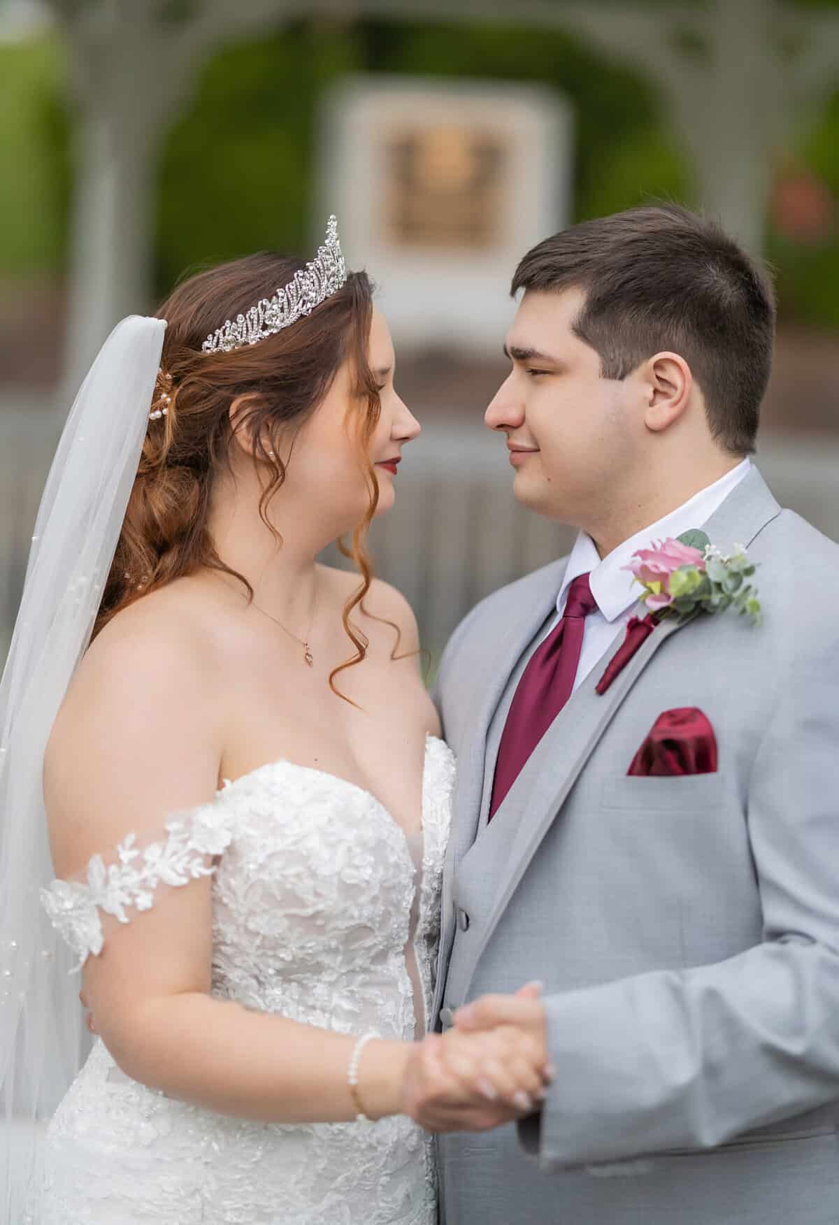 Wedding at the Springfield Elks lodge 37 A bride in a white lace gown and tiara and a groom in a gray suit with burgundy tie hold hands outdoors at their Springfield wedding, greenery in the background.