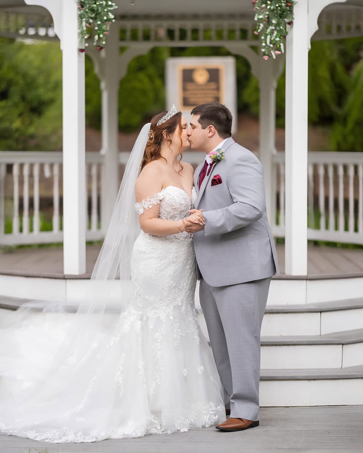 Wedding at the Springfield Elks lodge 39 A bride and groom in formal wedding attire share a kiss under a white gazebo at the Springfield Elks Lodge, decorated with greenery. The bride wears lace; the groom is in a light gray suit.