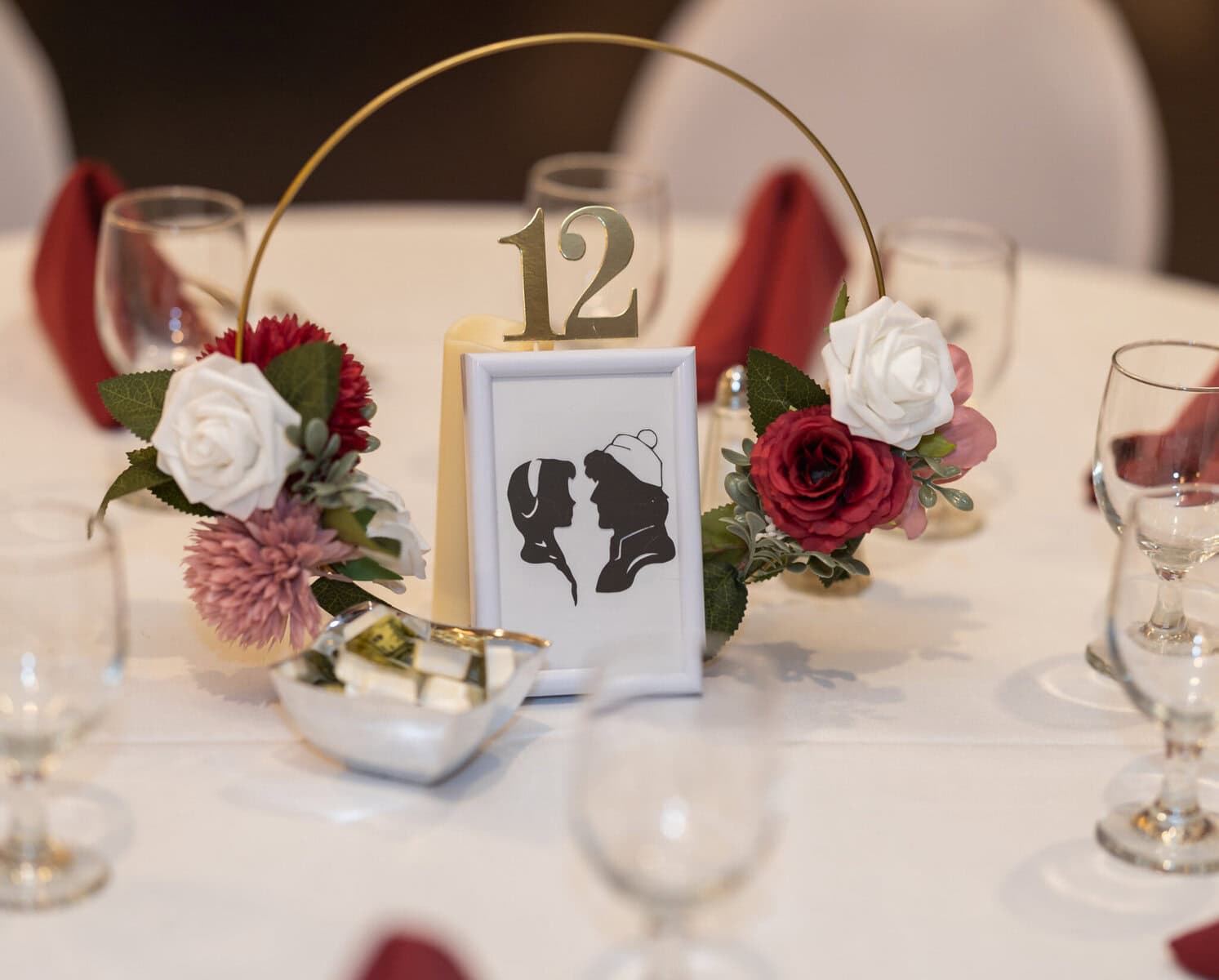 Wedding at the Springfield Elks lodge 49 A wedding table centerpiece at the Elks Lodge in Springfield features a gold “12” sign, a couple's silhouette, red and white flowers, and glasses on a white tablecloth.