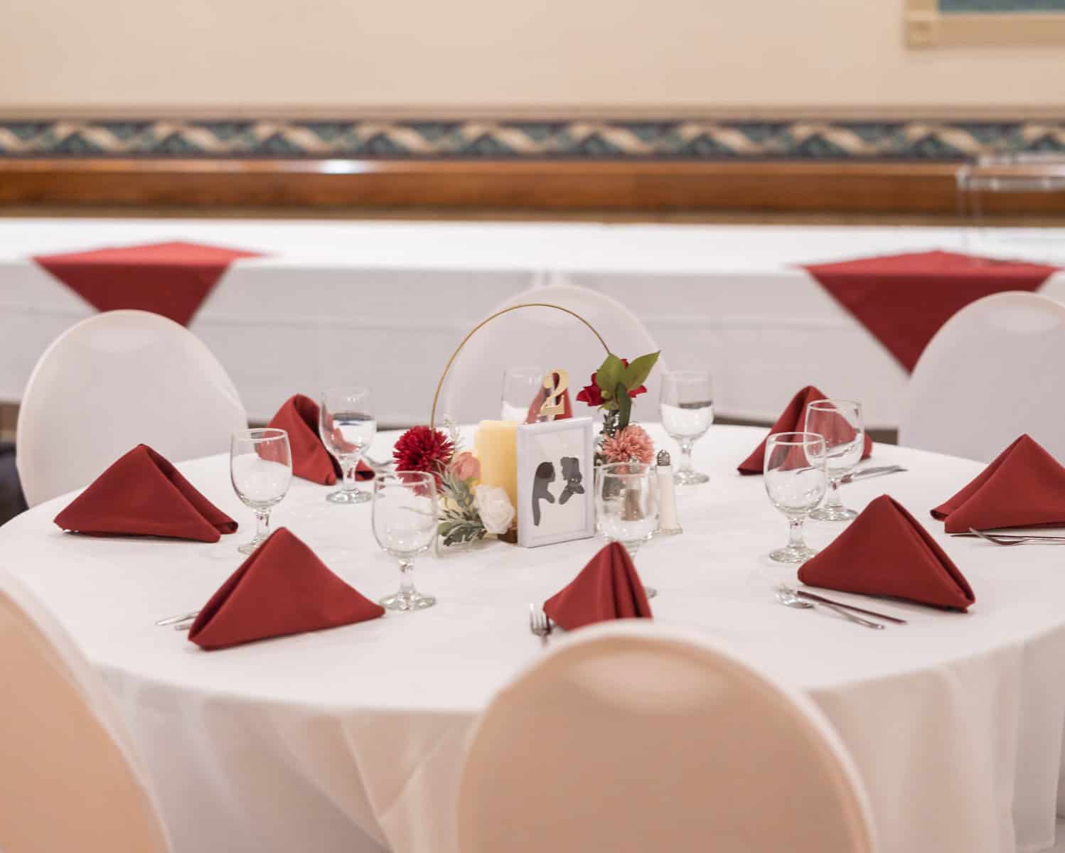 Wedding at the Springfield Elks lodge 50 A round banquet table at the Springfield Elks Lodge with white linen, burgundy napkins, floral centerpiece, and candles set for six at a wedding in a decorated event hall.