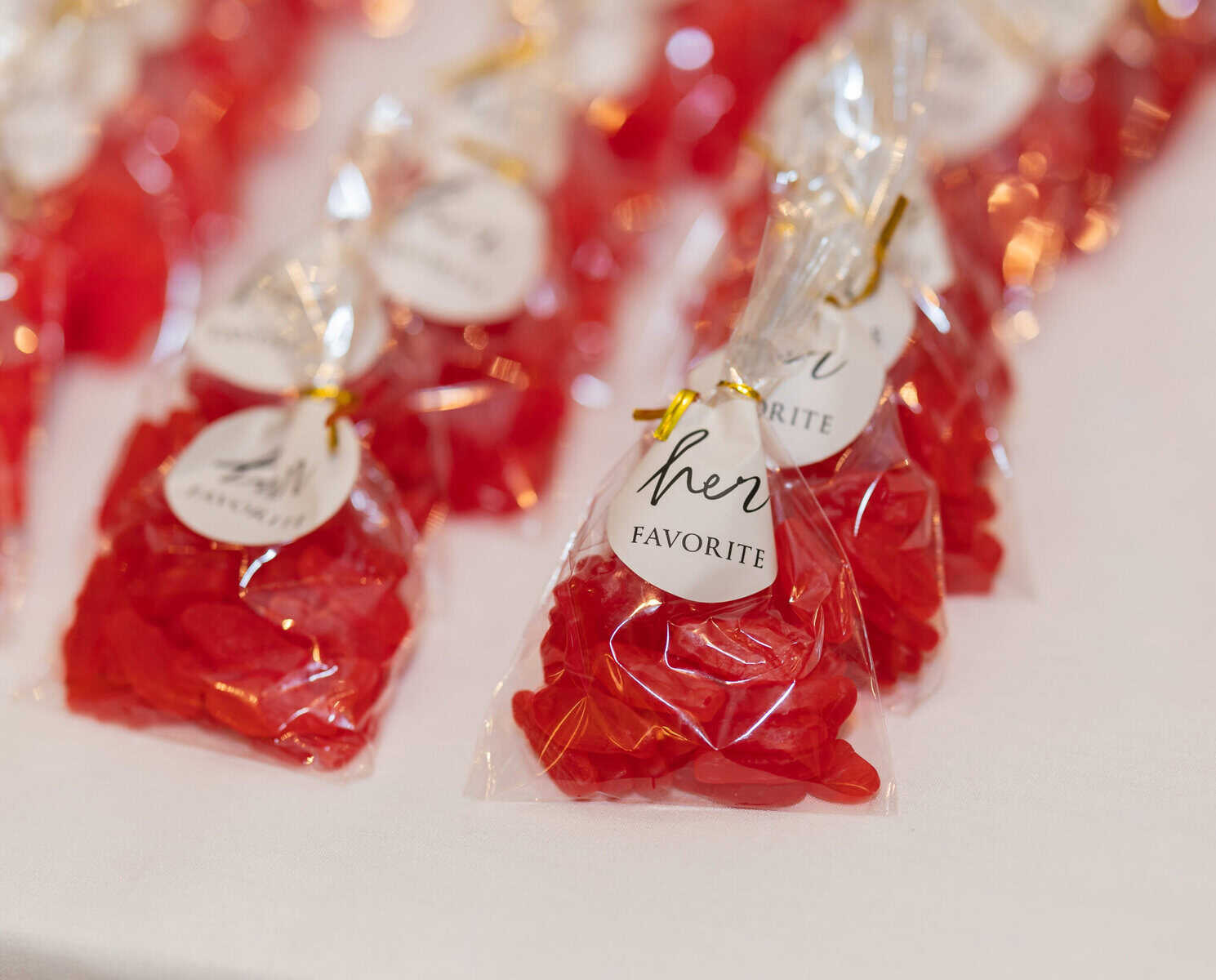 Wedding at the Springfield Elks lodge 51 Clear plastic bags of red gummy candies tied with gold twist ties, each labeled “her favorite,” arranged on a white tablecloth at a Springfield wedding at the Elks Lodge.