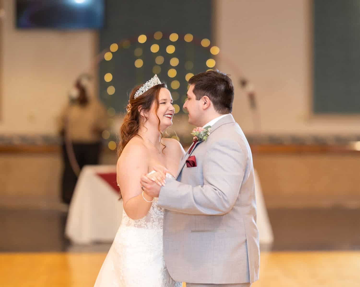 Wedding at the Springfield Elks lodge 60 A bride in a white gown and tiara smiles while dancing with the groom in a light gray suit at their Springfield wedding; warm fairy lights glow softly in the blurred background.