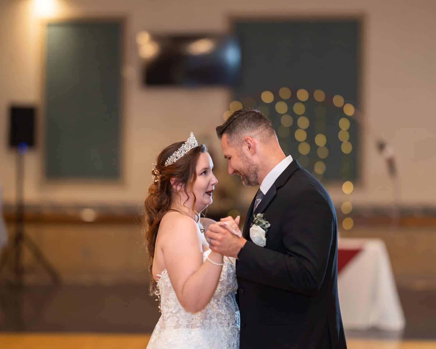 Wedding at the Springfield Elks lodge 62 A bride in a white gown and tiara and a groom in a black suit smile at each other while dancing indoors at their Springfield wedding reception at the Elks Lodge.