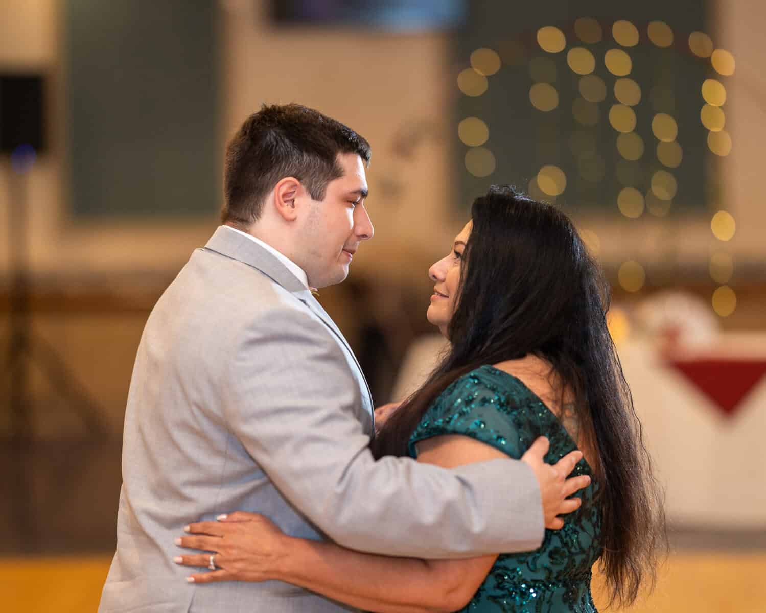 Wedding at the Springfield Elks lodge 65 A man in a light gray suit and a woman in a dark green dress embrace and smile, dancing at an Elks Lodge wedding in Springfield, with blurred lights glowing warmly in the background.