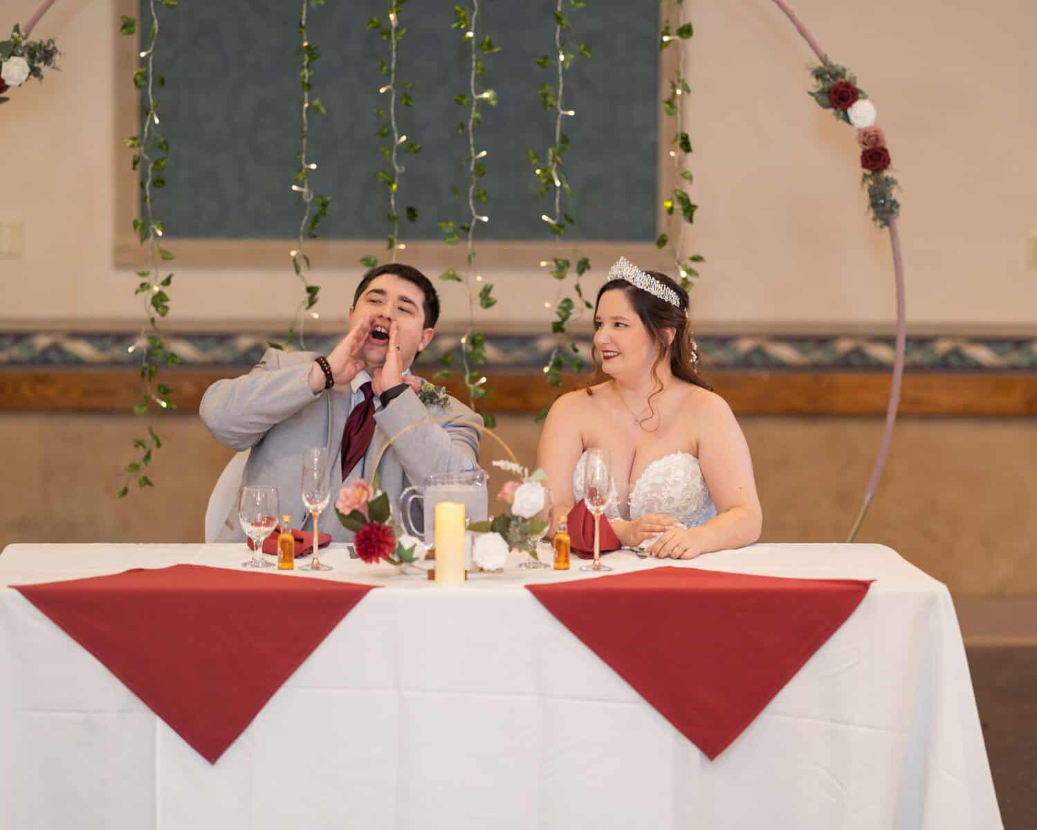 Wedding at the Springfield Elks lodge 66 A groom in a gray suit shouts playfully next to his smiling bride at a decorated Springfield Elks Lodge wedding table with red napkins and floral arrangements.
