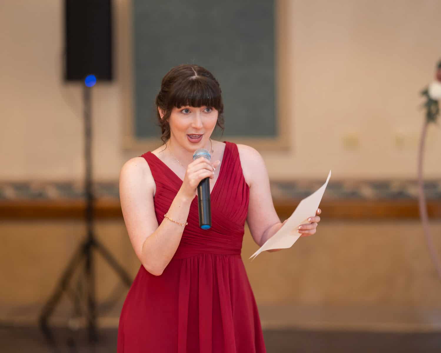 Wedding at the Springfield Elks lodge 67 A woman in a sleeveless red dress holds a microphone and a piece of paper, speaking at a formal wedding celebration indoors, possibly at the Springfield Elks Lodge.
