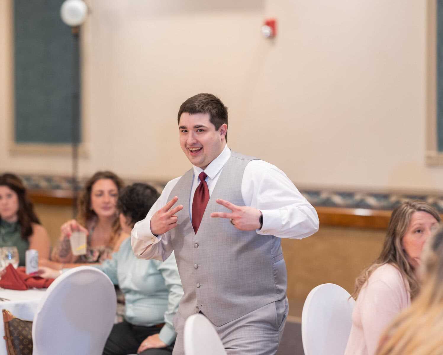 Wedding at the Springfield Elks lodge 68 A young man in a gray vest and red tie smiles and flashes peace signs with both hands at a wedding held at the Springfield Elks Lodge, with seated guests in the background.
