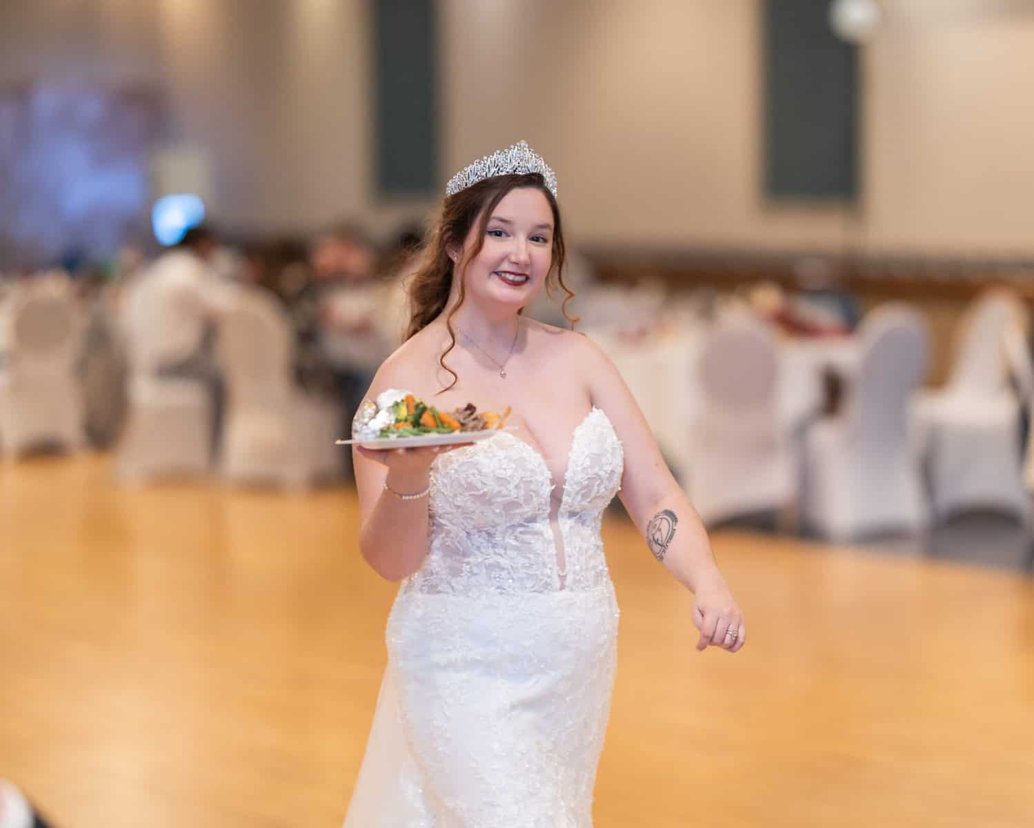 Wedding at the Springfield Elks lodge 69 A smiling bride in a white lace gown and tiara holds a plate of food while walking on a wooden floor at an Elks Lodge wedding reception in Springfield.