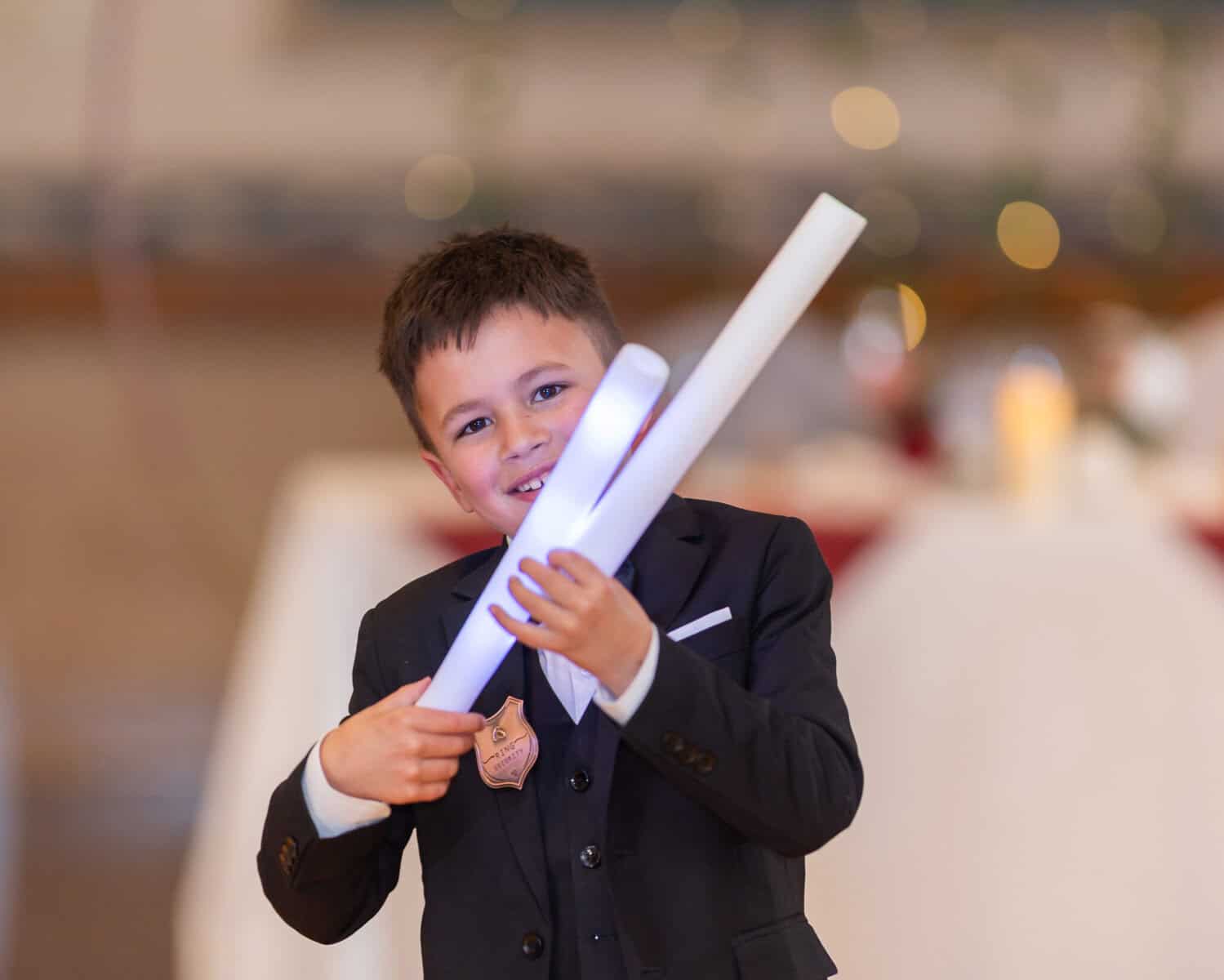 Wedding at the Springfield Elks lodge 73 A young boy in a black suit and bow tie smiles while holding two rolled-up white papers at a Springfield wedding. The softly blurred background shows lights and a decorated table.