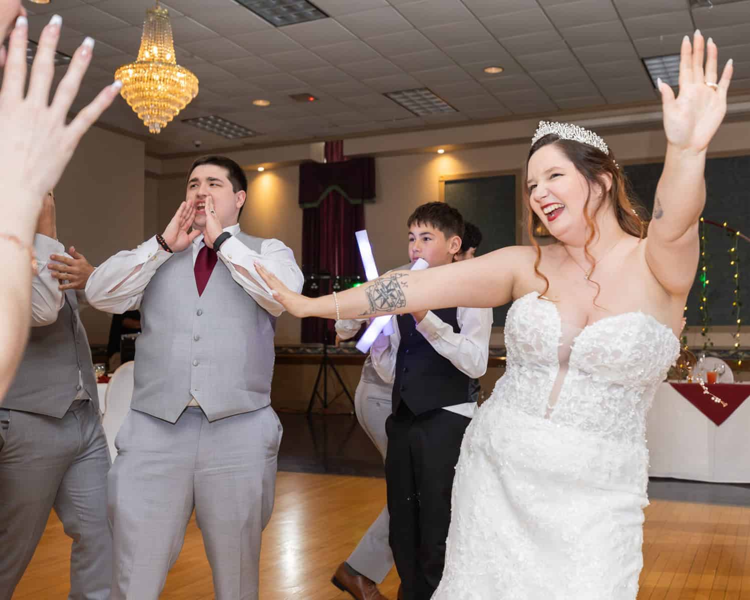 Wedding at the Springfield Elks lodge 76 A joyful bride in a strapless gown dances with arms raised as guests celebrate and cheer at a brightly lit Springfield Elks Lodge wedding reception.