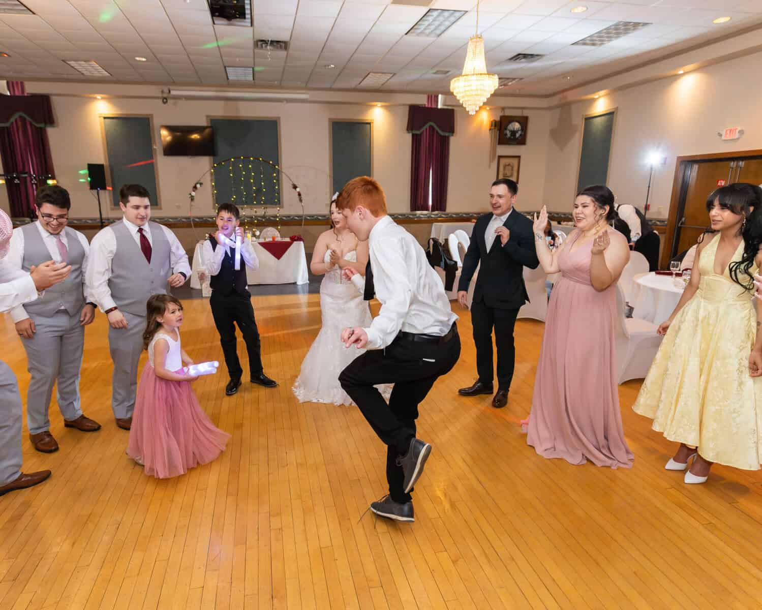 Wedding at the Springfield Elks lodge 78 A wedding reception at an Elks Lodge in Springfield, with guests dancing in a circle on polished wood as the bride twirls in her white dress among formal grey, pink, and yellow attire.