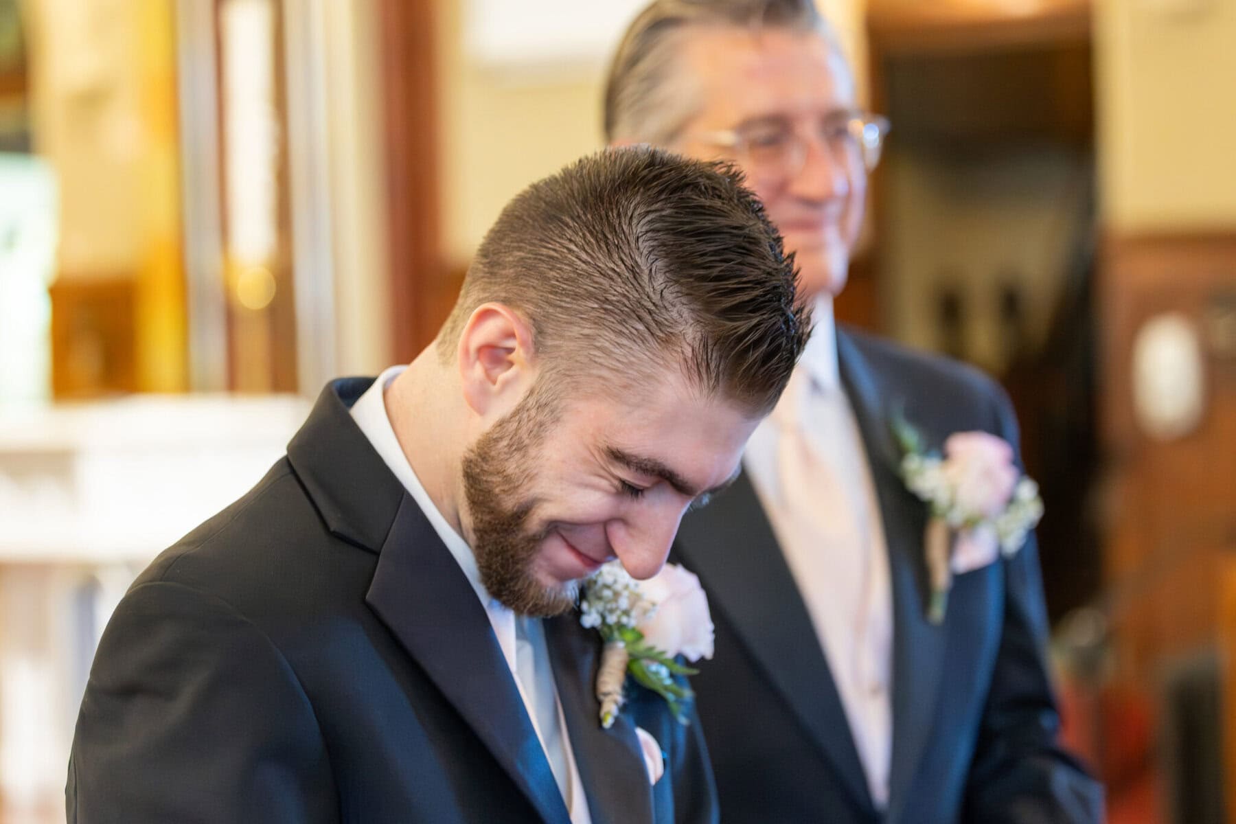 Wedding At St. Mary's Church in North Attleborough 24 A man in a suit smiles and looks down, with another man behind him—both wearing boutonnieres at a wedding in St. Mary’s Church.